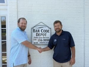 Gary and Cliff Hardesty shaking hands in front of the Bar Code Depot office and company sign, symbolizing the transition of leadership from father to son.